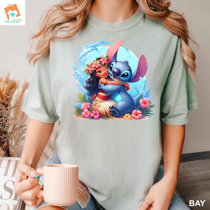 May include: A light blue t-shirt with a cartoon image of Stitch from Lilo & Stitch hugging a girl with brown hair and a flower lei. The girl is wearing a brown grass skirt and a pink flower behind her ear.