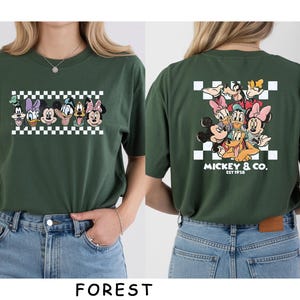 May include: A forest green t-shirt featuring a graphic of Disney characters on the front and back. The front shows a horizontal row of character faces over a checkerboard pattern. The back displays a larger group graphic with the text MICKEY & CO. EST 1928.