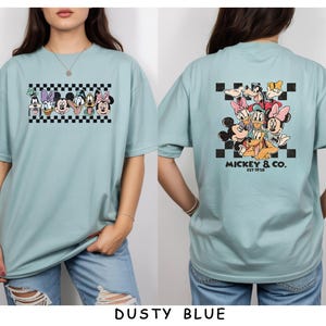 May include: A dusty blue t-shirt featuring a graphic print of Disney characters. The front displays a horizontal row of character faces over a black and white checkerboard pattern. The back shows a larger group of characters with the text MICKEY & CO. EST 1928.