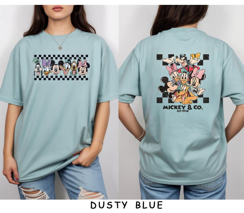 May include: A dusty blue t-shirt featuring a graphic print of Disney characters. The front displays a horizontal row of character faces over a black and white checkerboard pattern. The back shows a larger group of characters with the text MICKEY & CO. EST 1928.
