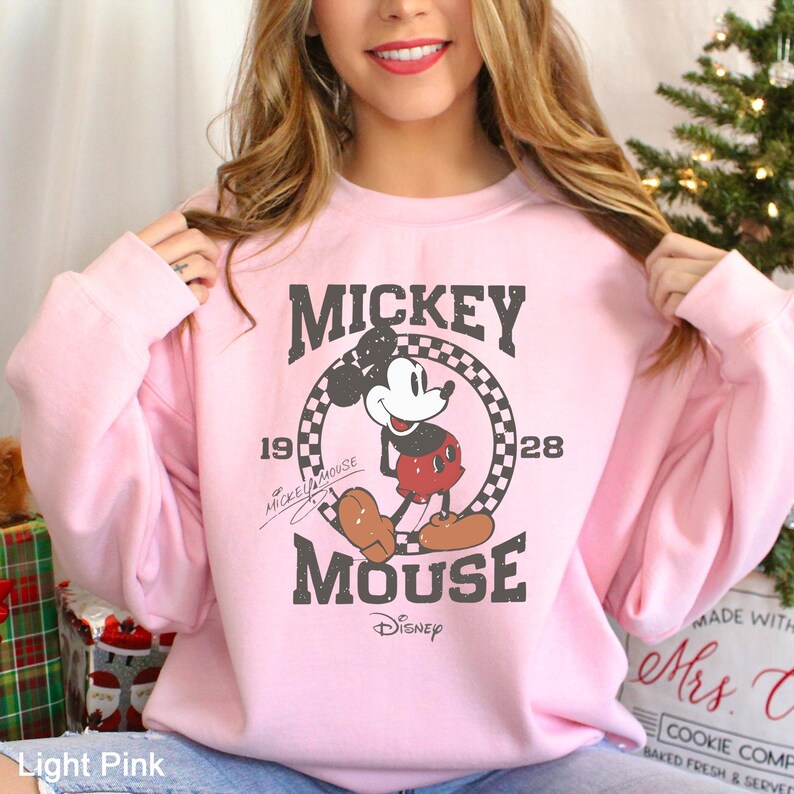 Puede incluir: Una sudadera rosa claro con un gr&aacute;fico vintage de Mickey Mouse. El gr&aacute;fico presenta a Mickey Mouse con sus caracter&iacute;sticos pantalones cortos rojos y zapatos amarillos, de pie en un c&iacute;rculo a cuadros blanco y negro con el texto "Mickey Mouse" y "1928" arriba y abajo del c&iacute;rculo. El texto "Mickey Mouse" est&aacute; escrito en una fuente manuscrita.