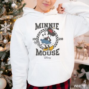 Puede incluir: Sudadera con capucha blanca con un gr&aacute;fico de estilo vintage de Minnie Mouse. El gr&aacute;fico presenta a Minnie Mouse con su caracter&iacute;stico vestido rojo y lazo de lunares, con el texto "Minnie Mouse" y "1928" en una fuente retro.