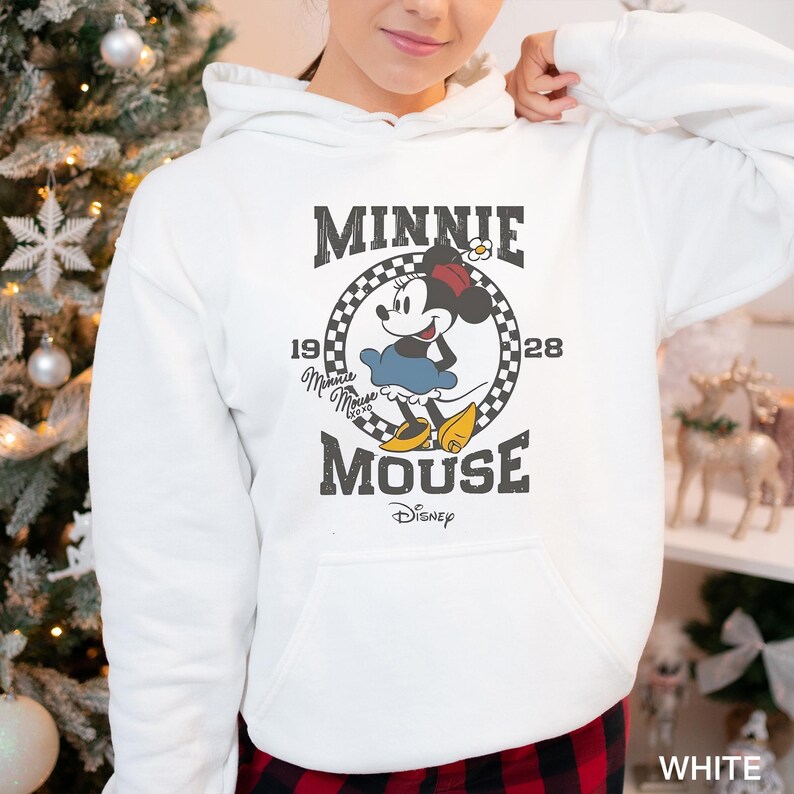 Puede incluir: Sudadera con capucha blanca con un gr&aacute;fico de estilo vintage de Minnie Mouse. El gr&aacute;fico presenta a Minnie Mouse con su caracter&iacute;stico vestido rojo y lazo de lunares, con el texto "Minnie Mouse" y "1928" en una fuente retro.