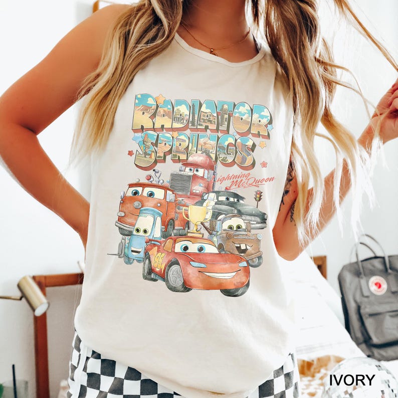 Puede incluir: Camiseta sin mangas color marfil con un gr&aacute;fico colorido de personajes de la pel&iacute;cula Cars, incluido Rayo McQueen. El texto "Radiator Springs" se muestra encima de los personajes en una fuente retro.