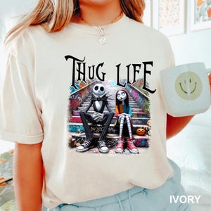 Puede incluir: Una camiseta blanca con un estampado gr&aacute;fico de Jack Skellington y Sally de Pesadilla antes de Navidad sentados en una escalera cubierta de grafiti. El texto "THUG LIFE" est&aacute; impreso encima de ellos.