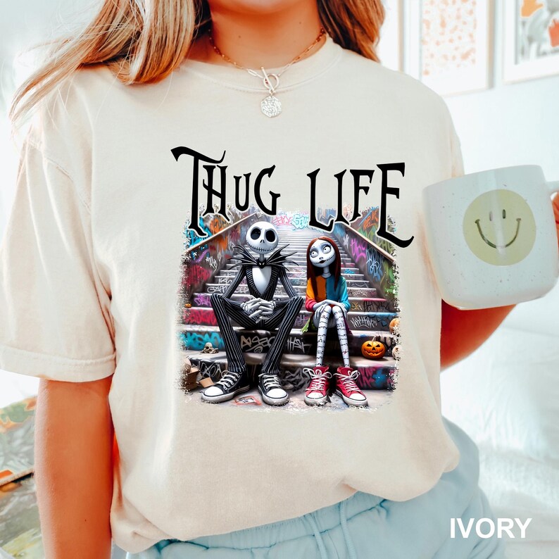 Puede incluir: Una camiseta blanca con un estampado gr&aacute;fico de Jack Skellington y Sally de Pesadilla antes de Navidad sentados en una escalera cubierta de grafiti. El texto "THUG LIFE" est&aacute; impreso encima de ellos.