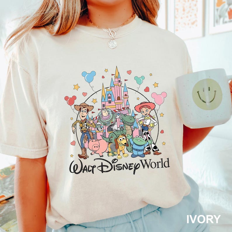 Puede incluir: Camiseta color marfil con un gr&aacute;fico colorido de los personajes de Toy Story frente a un castillo, con las palabras "Walt Disney World" debajo. Tambi&eacute;n se ve una taza blanca con una carita sonriente.