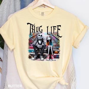 Puede incluir: Una camiseta amarilla con un estampado gr&aacute;fico de Jack Skellington y Sally de Pesadilla antes de Navidad sentados en un tramo de escaleras. El texto "Thug Life" est&aacute; impreso encima de ellos.