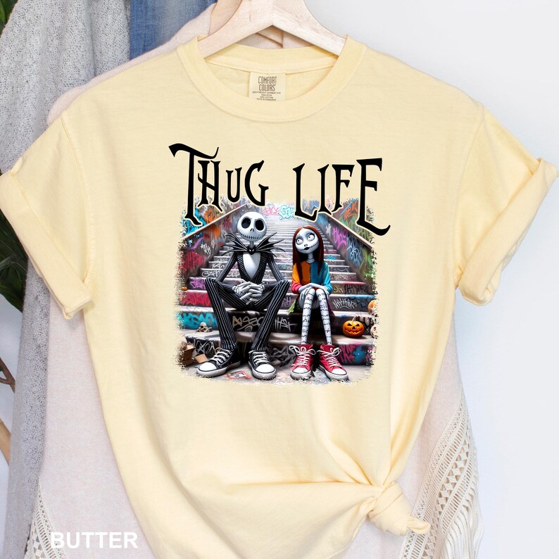 Puede incluir: Una camiseta amarilla con un estampado gr&aacute;fico de Jack Skellington y Sally de Pesadilla antes de Navidad sentados en un tramo de escaleras. El texto "Thug Life" est&aacute; impreso encima de ellos.
