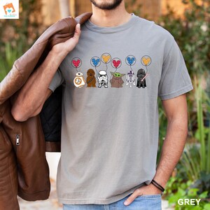May include: A grey t-shirt with a graphic of Star Wars characters holding balloons shaped like Mickey Mouse ears. The characters include Chewbacca, Stormtrooper, Yoda, R2-D2, and Darth Vader. The balloons are red, blue, yellow, and green.