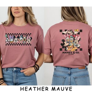 May include: A heather mauve t-shirt featuring a front graphic of Disney characters in a row above a black and white checkerboard pattern and a back graphic of the same characters clustered together with the text Mickey & Co. Est 1928.