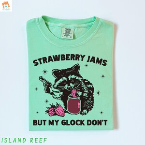 May include: A light green t-shirt with a black and white illustration of a raccoon holding a jar of strawberry jam and a gun. The text "Strawberry Jams But My Glock Don't" is printed on the shirt.