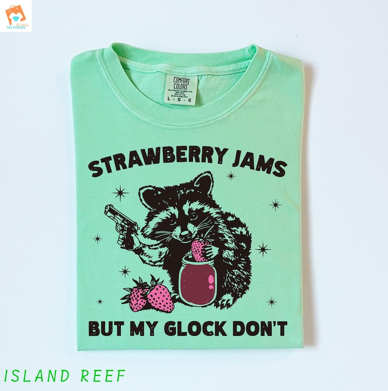 May include: A light green t-shirt with a black and white illustration of a raccoon holding a jar of strawberry jam and a gun. The text "Strawberry Jams But My Glock Don't" is printed on the shirt.