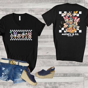 May include: A black V-neck t-shirt featuring a graphic of Disney characters over a white and black checkered pattern on the front and back. The back design includes the text MICKEY & CO. EST 1928. Also includes blue denim shorts and navy wedge sandals.