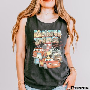 Puede incluir: Camiseta sin mangas negra con un gr&aacute;fico colorido de personajes de la pel&iacute;cula Cars, incluyendo a Rayo McQueen y el texto "Radiator Springs". El dise&ntilde;o tiene un estilo vintage y retro.