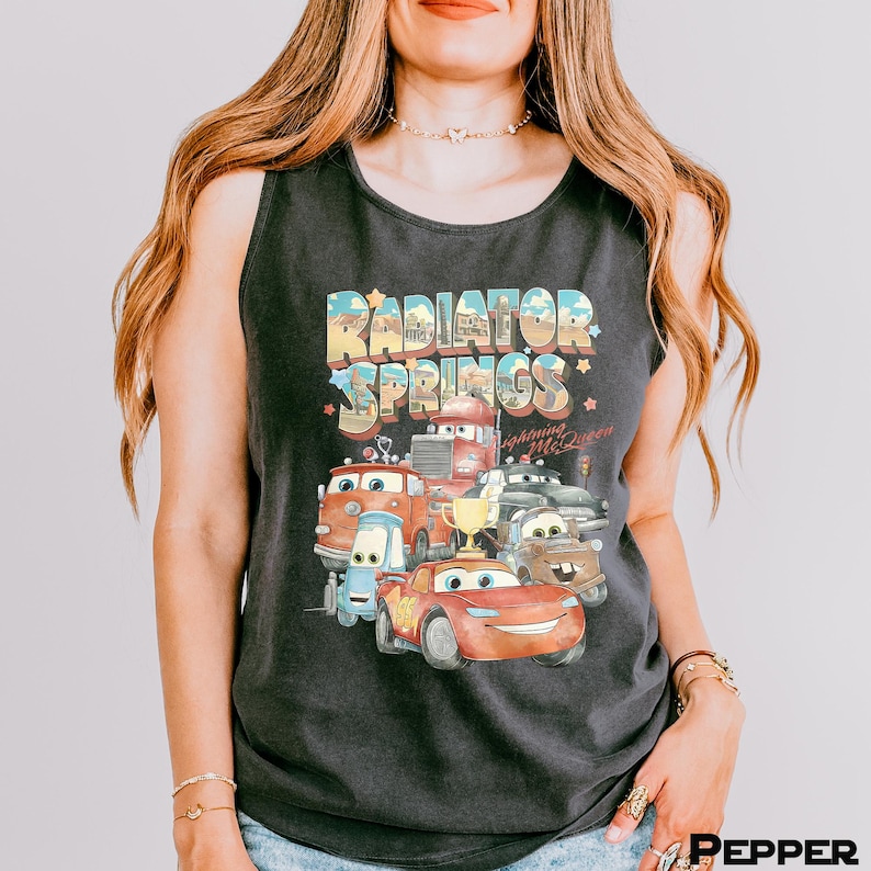 Puede incluir: Camiseta sin mangas negra con un gr&aacute;fico colorido de personajes de la pel&iacute;cula Cars, incluyendo a Rayo McQueen y el texto "Radiator Springs". El dise&ntilde;o tiene un estilo vintage y retro.
