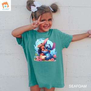 May include: A teal blue t-shirt with a graphic of Stitch from Lilo & Stitch hugging a girl with a flower lei. The girl is wearing a pink and white dress and has brown hair. The graphic is surrounded by blue waves and pink flowers.