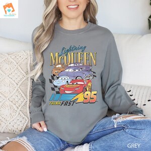 May include: A grey long-sleeved sweatshirt featuring a graphic of Lightning McQueen and other Cars characters. The text "Lightning McQueen" and "Think Fast" are printed on the shirt. The number "95" is also printed on the shirt.