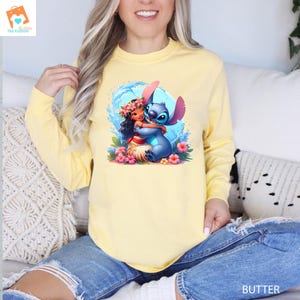 May include: A yellow long-sleeved sweatshirt with a graphic print of Stitch from Lilo & Stitch hugging Moana from Moana. The graphic is colorful and features a blue ocean wave background.