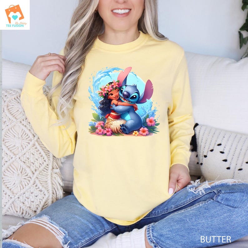 May include: A yellow long-sleeved sweatshirt with a graphic print of Stitch from Lilo & Stitch hugging Moana from Moana. The graphic is colorful and features a blue ocean wave background.