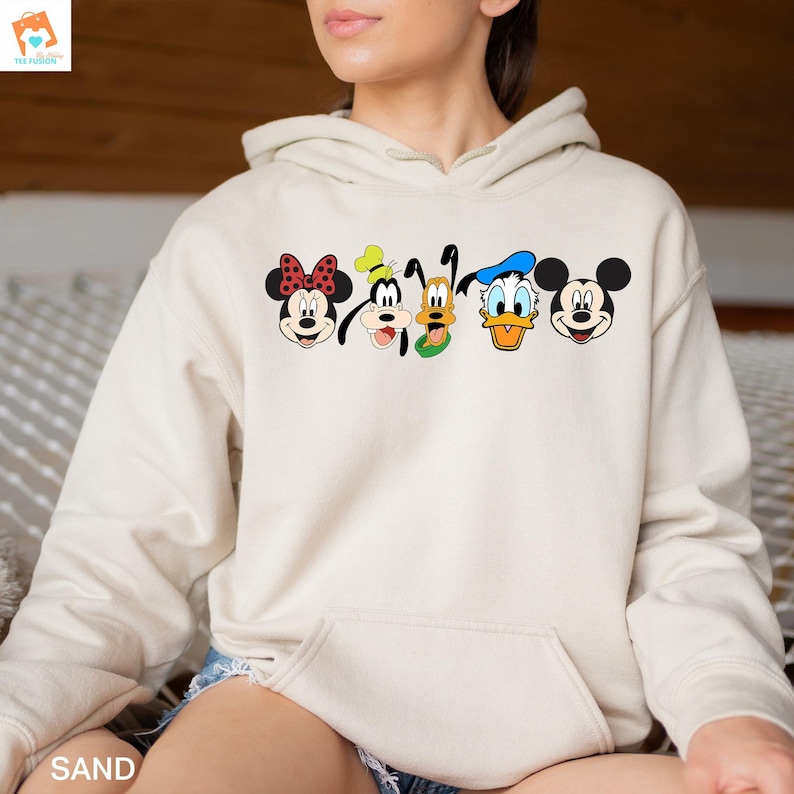 Op de afbeelding: Een cr&egrave;mekleurige hoodie met een afbeelding van Mickey Mouse, Minnie Mouse, Donald Duck, Goofy en Pluto. De personages staan in een rij, met hun gezichten naar voren gericht. De hoodie heeft een capuchon met trekkoord en een kangoeroezak.