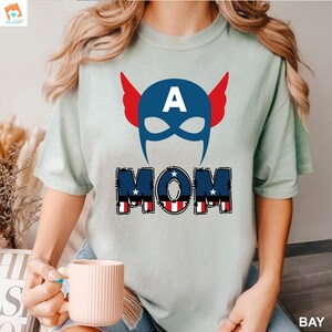 May include: A light blue t-shirt with a red, white, and blue graphic of a superhero mask with the letter "A" on the forehead and the word "MOM" underneath. The word "MOM" is in a red, white, and blue patchwork style.