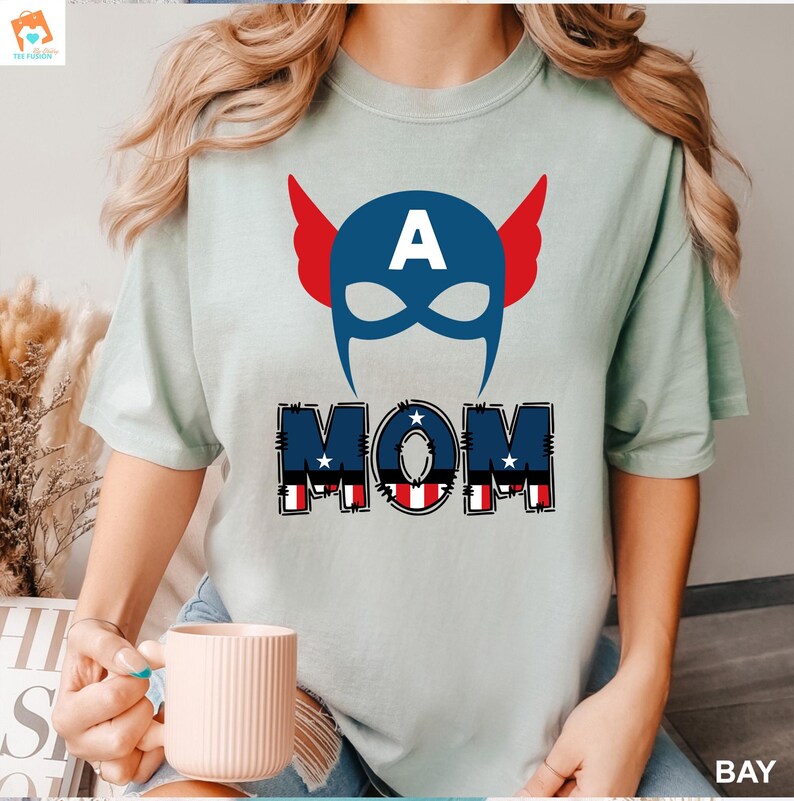 May include: A light blue t-shirt with a red, white, and blue graphic of a superhero mask with the letter "A" on the forehead and the word "MOM" underneath. The word "MOM" is in a red, white, and blue patchwork style.