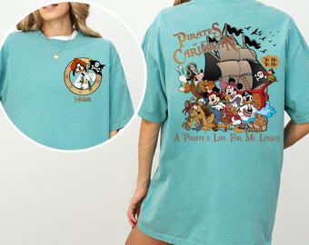 Könnte beinhalten: Ein türkisfarbenes T-Shirt mit einem grafischen Aufdruck von Mickey Mouse und anderen Disney-Charakteren, die als Piraten auf einem Schiff verkleidet sind. Der Text "Pirates of the Caribbean" und "A Pirate's Life For Me Lyrics" ist auf dem Shirt aufgedruckt.