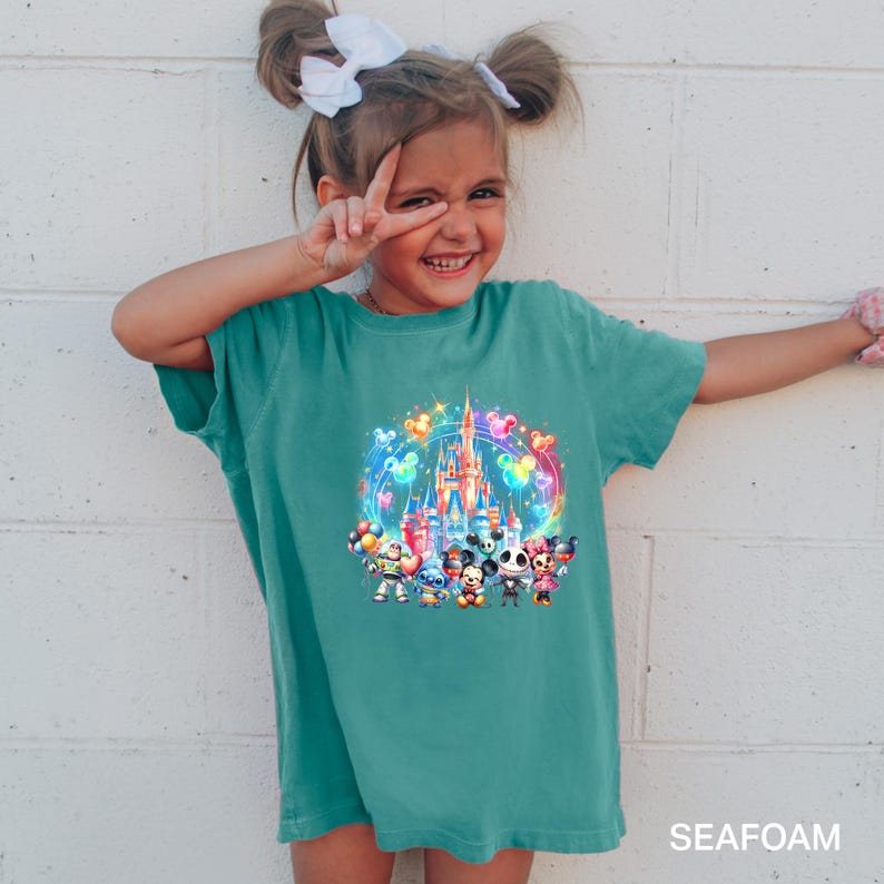 Puede incluir: Una ni&ntilde;a peque&ntilde;a lleva una camiseta azul turquesa con un gr&aacute;fico colorido de personajes de Disney frente al Castillo de Cenicienta. El gr&aacute;fico incluye a Mickey Mouse, Minnie Mouse, Donald Duck, Goofy, Pluto y otros personajes de Disney. La camiseta tiene la palabra "SEAFOM" impresa en la esquina inferior derecha.