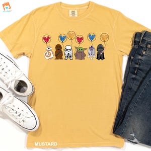 May include: A yellow t-shirt with a graphic of Star Wars characters holding balloons shaped like Mickey Mouse heads. The characters include R2-D2, Chewbacca, Darth Vader, a Stormtrooper, and Baby Yoda.