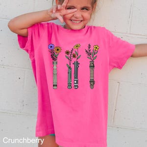 Puede incluir: Una camiseta rosa con un gr&aacute;fico de cuatro sables de luz con flores que crecen de ellos. Los sables de luz son verde, azul, morado y rojo.