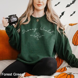 May include: Forest green sweatshirt with the text "Disney World" in white script, along with a Mickey Mouse graphic and the year "1971". The sweatshirt is a solid color and has a crew neck. A black mug with the text "Witches Brew" is held in the left hand.