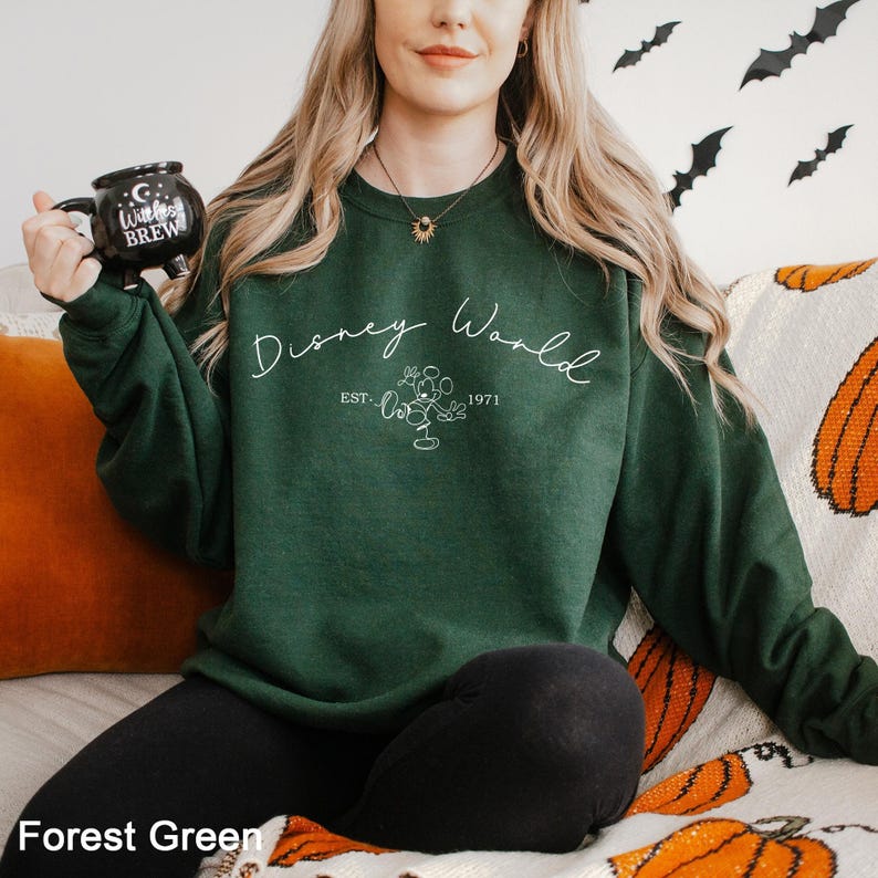 May include: Forest green sweatshirt with the text "Disney World" in white script, along with a Mickey Mouse graphic and the year "1971". The sweatshirt is a solid color and has a crew neck. A black mug with the text "Witches Brew" is held in the left hand.