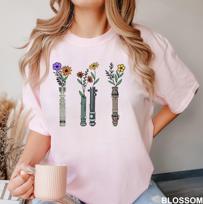 Puede incluir: Camiseta rosa con un gr&aacute;fico de cuatro sables de luz con flores en los mangos. Los sables de luz son verde, azul, morado y rojo.
