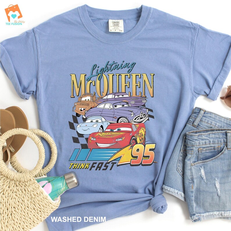 May include: A light blue t-shirt featuring a vintage-style graphic of Lightning McQueen and other Cars characters. The graphic includes the text "Lightning McQueen" and "Think Fast 95".