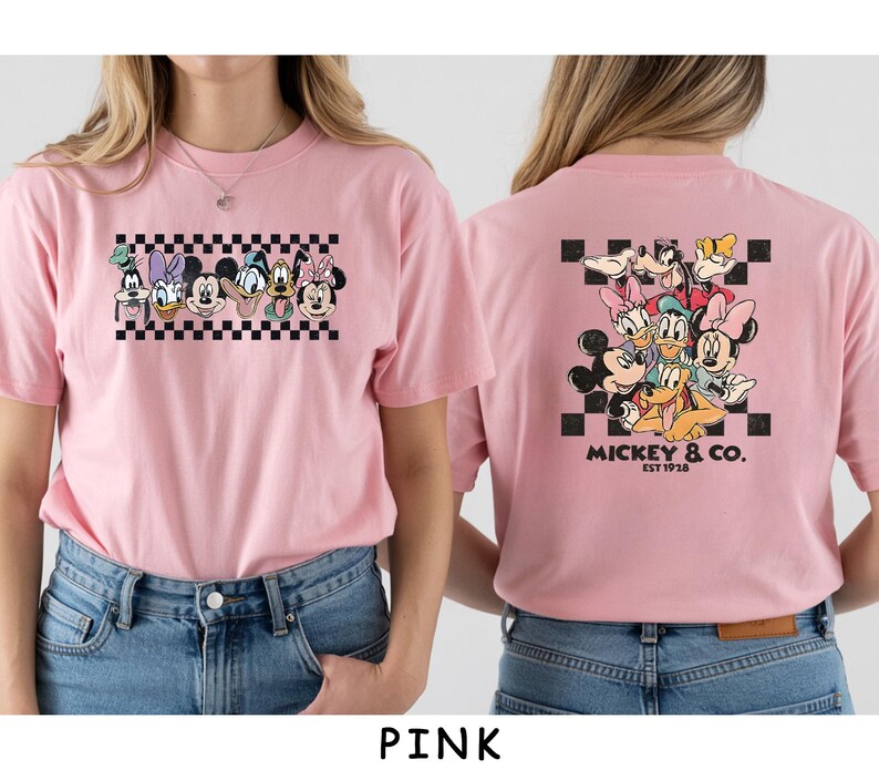 May include: A light pink t-shirt featuring a graphic of Disney characters on the front and back. The front shows a horizontal row of character faces over a black and white checkerboard pattern. The back displays a larger group of characters.