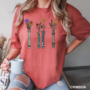 Puede incluir: Una camiseta roja con un gr&aacute;fico de cuatro sables de luz con flores que crecen desde la parte superior. Los sables de luz son verde, azul, rojo y amarillo.