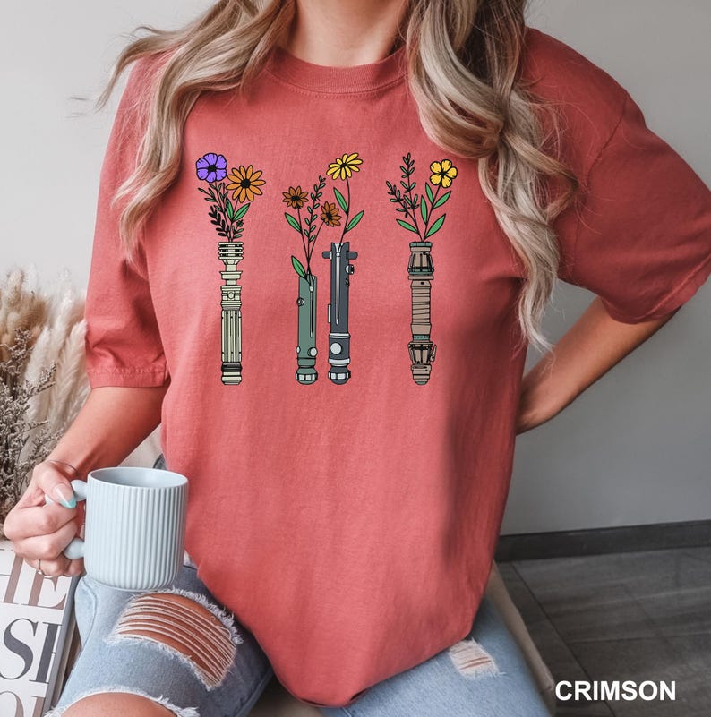 Puede incluir: Una camiseta roja con un gr&aacute;fico de cuatro sables de luz con flores que crecen desde la parte superior. Los sables de luz son verde, azul, rojo y amarillo.