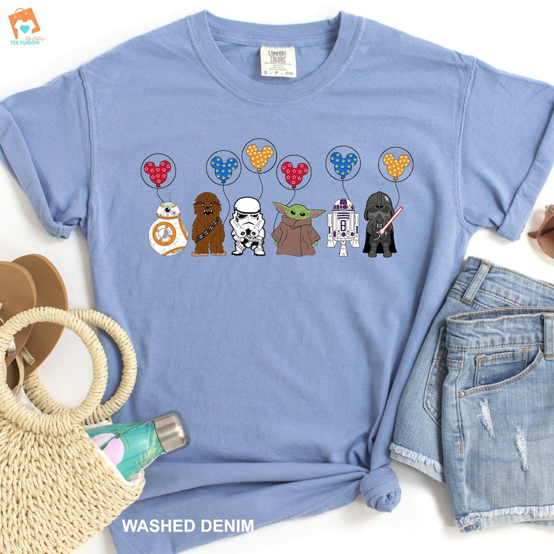 May include: A light blue t-shirt with a graphic of Star Wars characters holding Mickey Mouse balloons. The characters include Chewbacca, a Stormtrooper, Yoda, R2-D2, and Darth Vader.