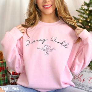 May include: Light pink sweatshirt with the text "Disney World" in a script font, along with a Mickey Mouse graphic and the year "1971". The sweatshirt is a solid light pink color.