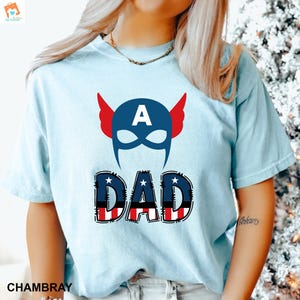 May include: A light blue t-shirt with a red, white, and blue graphic of a superhero mask with the letter "A" on the forehead and the word "DAD" below it.