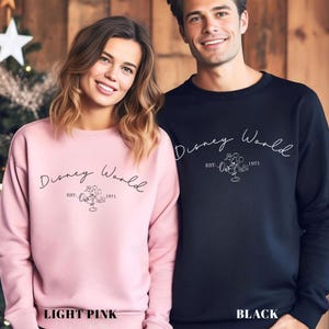 May include: Light pink and black sweatshirts with the text "Disney World" in a script font. Each sweatshirt has a Mickey Mouse graphic and the text "EST. 1971". The bottom of the image has the text "LIGHT PINK" and "BLACK".