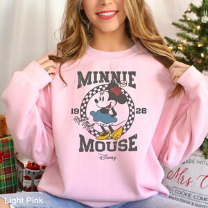 Puede incluir: Una sudadera rosa claro con un gr&aacute;fico de estilo vintage de Minnie Mouse. El gr&aacute;fico presenta a Minnie Mouse con su caracter&iacute;stico vestido rojo de lunares y lazo, con el texto "Minnie Mouse" y "Disney" debajo.