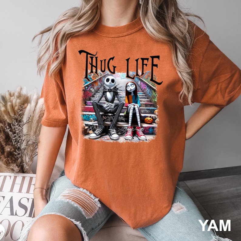 Puede incluir: Camiseta naranja con un estampado gr&aacute;fico de Jack Skellington y Sally de Pesadilla Antes de Navidad sentados en un tramo de escaleras con grafitis. El texto "THUG LIFE" est&aacute; impreso sobre los personajes.