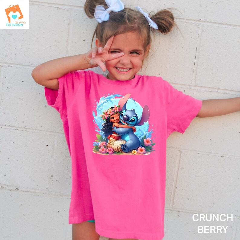 May include: A pink t-shirt with a graphic of Stitch from Lilo & Stitch hugging Moana from Moana. The graphic is set against a blue wave background with pink flowers.