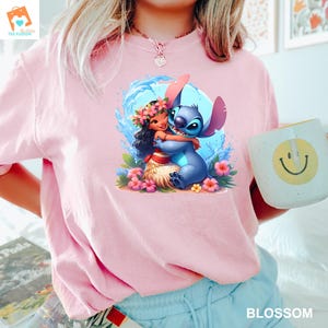 May include: Pink t-shirt with a cartoon image of Stitch from Lilo & Stitch hugging a girl with brown hair and a flower crown. The girl is wearing a grass skirt and holding a flower.