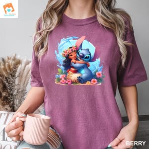 May include: A purple t-shirt with a cartoon image of Stitch and Lilo from the Disney movie Lilo & Stitch. Stitch is blue and has a big smile. Lilo is wearing a flower lei and has brown hair. The image is surrounded by flowers and water.