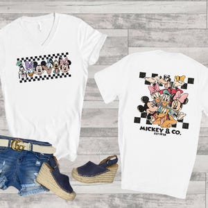 May include: A white V-neck t-shirt featuring a graphic of Disney characters on the front and back. The front shows a horizontal row of character faces over a black and white checkerboard pattern. The back displays a larger group of characters.