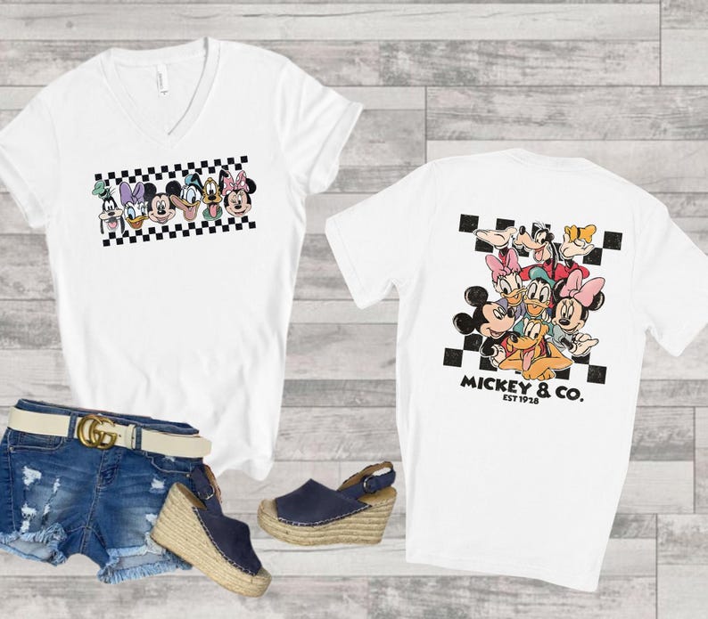 May include: A white V-neck t-shirt featuring a graphic of Disney characters on the front and back. The front shows a horizontal row of character faces over a black and white checkerboard pattern. The back displays a larger group of characters.