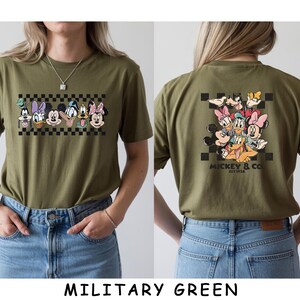 May include: An olive green t-shirt featuring a graphic of Disney characters including Mickey Mouse, Minnie Mouse, Donald Duck, Daisy Duck, Goofy, and Pluto. The front displays the characters in a row over a black and white checkerboard pattern.